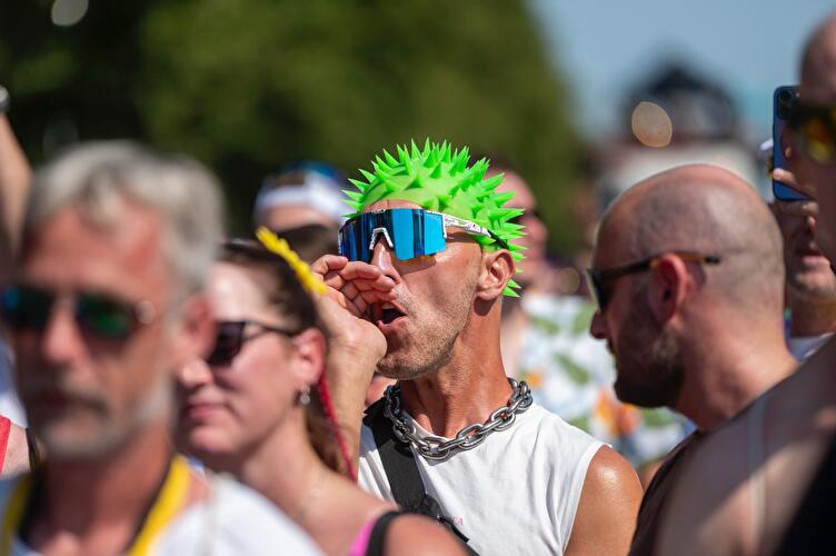 Nahaufnahme eines Mannes mit grüner Stachel-Perücke, verspiegelter Sonnenbrille und Kettenkette bei der Rave the Planet Parade, der lautstark ruft, umgeben von anderen Zuschauer:innen.