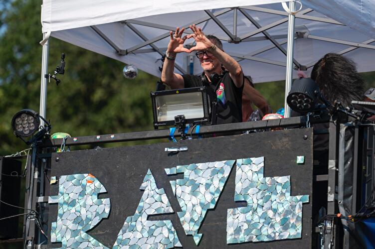 Der DJ Dr. Motte auf einem Truck der Rave the Planet Parade formt mit seinen Händen ein Herz über dem mit CD-Mosaik verzierten "Rave"-Schriftzug, umgeben von Scheinwerfern und einer Discokugel.