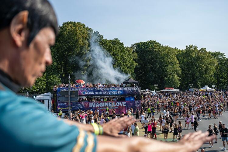 Dichte Menschenmenge auf der Straße des 17. Juni bei der Rave the Planet Parade in Berlin, umgeben von Bäumen, mit Musik-Trucks und Blick auf das Brandenburger Tor im Hintergrund.