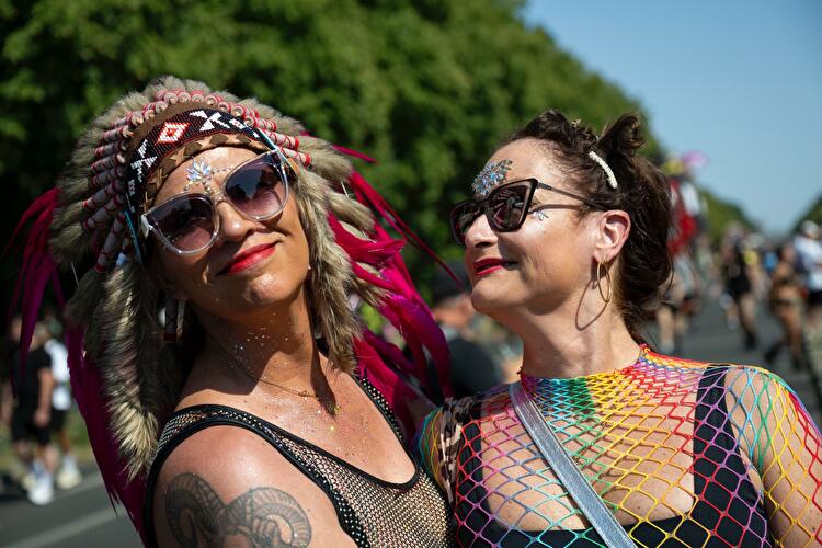 Zwei Frauen bei der Rave the Planet Parade blicken sich lächelnd an, eine mit Federkopfschmuck und Glitzer-Make-up, die andere mit buntem Netzoberteil und Strasssteinen im Gesicht, im Hintergrund Bäume und weitere Paradeteilnehmer:innen.