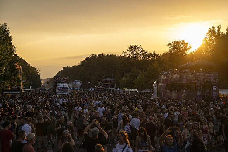 Menschenmenge und Musik-Trucks auf der Straße des 17. Juni bei Sonnenuntergang während der Rave the Planet Parade in Berlin, im Hintergrund die Siegessäule unter goldenem Abendhimmel.