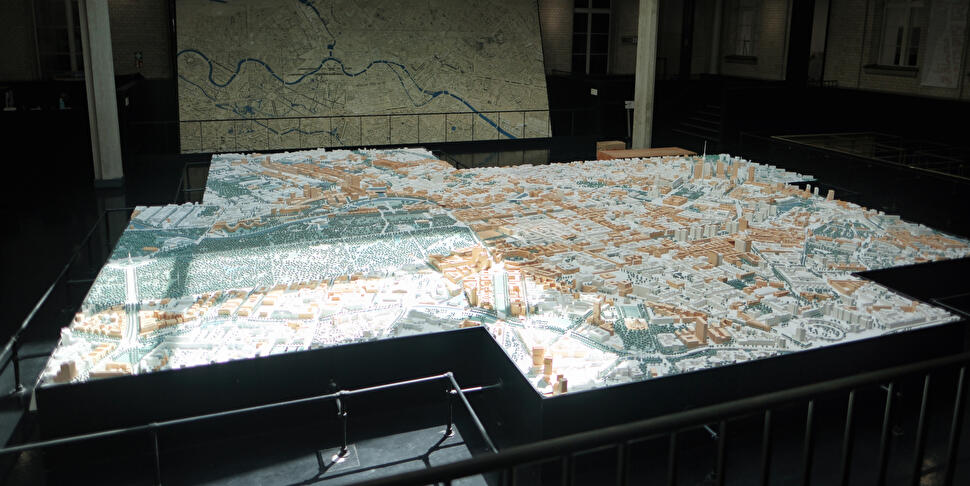 Großes Stadtmodell von Berlin in einer Ausstellungshalle, im Hintergrund eine historische Stadtplan-Wandkarte.