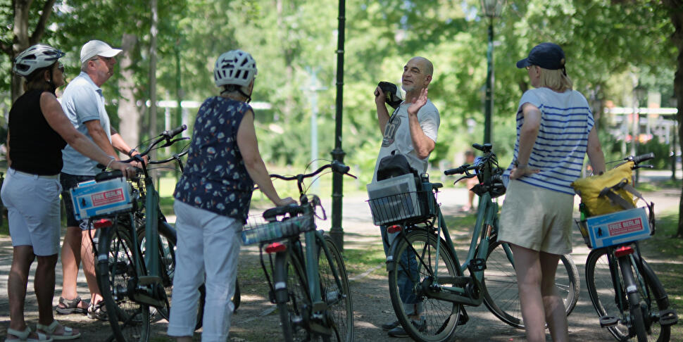 Eine Radtourgruppe mit "Free Berlin"-Fahrrädern steht auf einem schattigen Parkweg und hört einem Guide zu.
