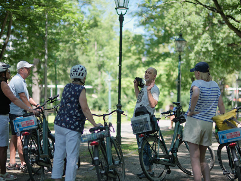 Eine Radtourgruppe mit "Free Berlin"-Fahrrädern steht auf einem schattigen Parkweg und hört einem Guide zu.