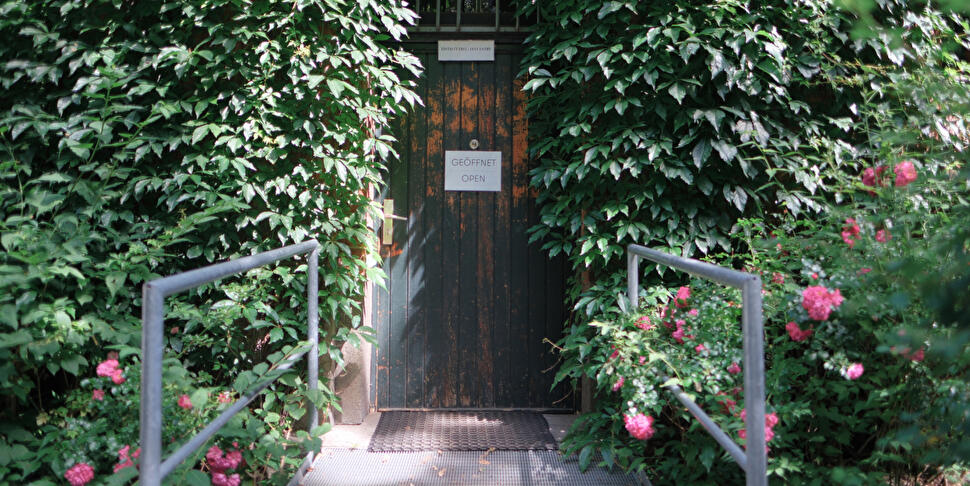 Efeu- und rosenumrankter Eingang mit dunkler Holztür und Schild "Geöffnet / Open", davor eine Metallrampe mit Geländer.