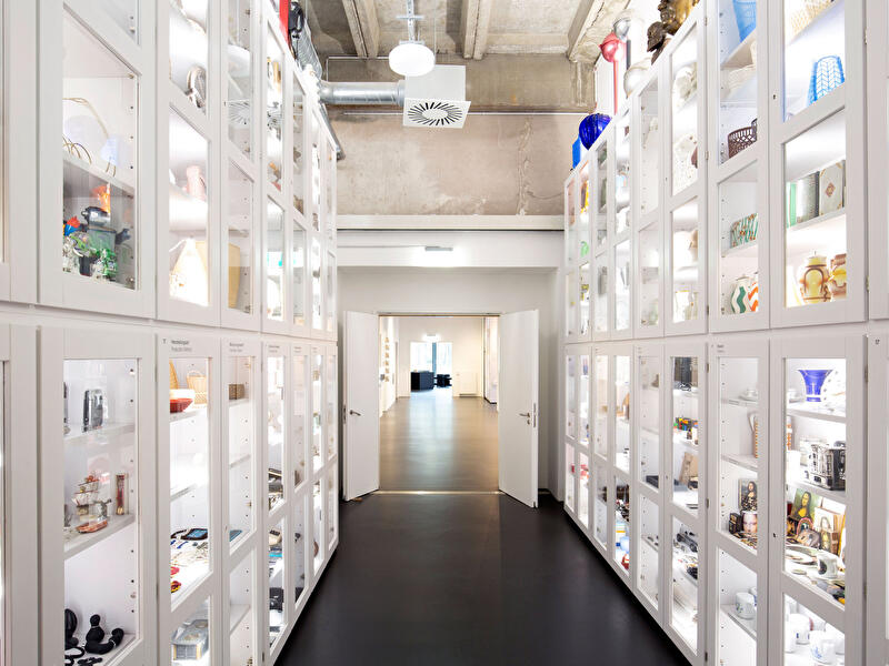 Gang mit beleuchteten Vitrinen voller Design- und Alltagsobjekte im Museumsdepot.
