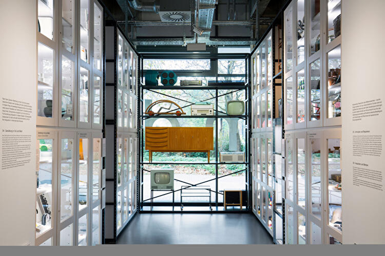 Ausstellungsraum mit Vitrinen und historischen Designobjekten vor großer Fensterfront.