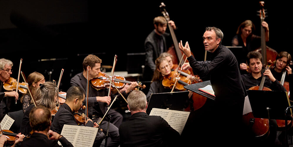 Dirigent leitet Orchester während eines klassischen Konzerts auf beleuchteter Bühne.
