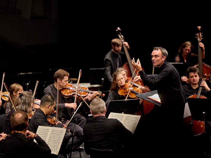 Dirigent leitet Orchester während eines klassischen Konzerts auf beleuchteter Bühne.