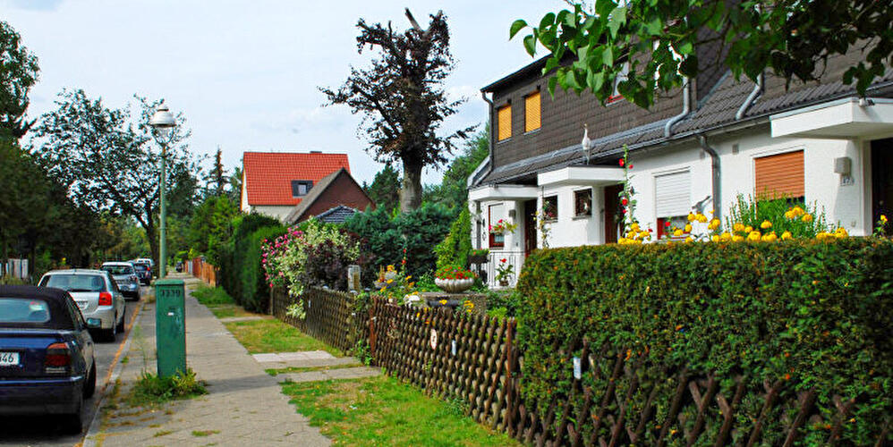 Wohnstraße mit parkenden Autos sowie Reihenhäusern mit gepflegten Vorgärten, Hecken und Blumen entlang des Gehwegs.