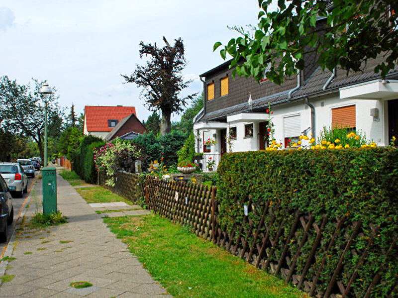 Wohnstraße mit parkenden Autos sowie Reihenhäusern mit gepflegten Vorgärten, Hecken und Blumen entlang des Gehwegs.