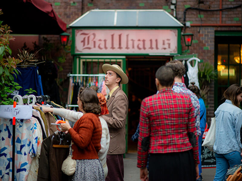 Menschen stöbern an einem Secondhand-Kleiderstand vor einem Gebäude mit der Aufschrift „Ballhaus“. Ein Schild am Kleiderständer weist auf einen Preis von „alles 5 €“ hin.