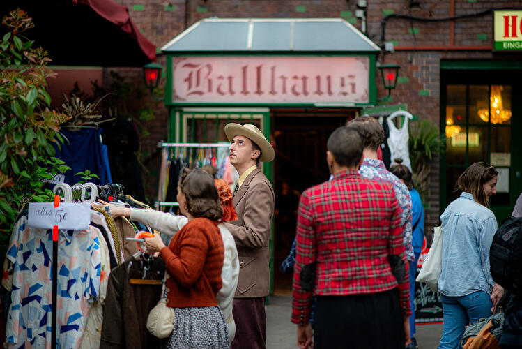 Menschen stöbern an einem Secondhand-Kleiderstand vor einem Gebäude mit der Aufschrift „Ballhaus“.