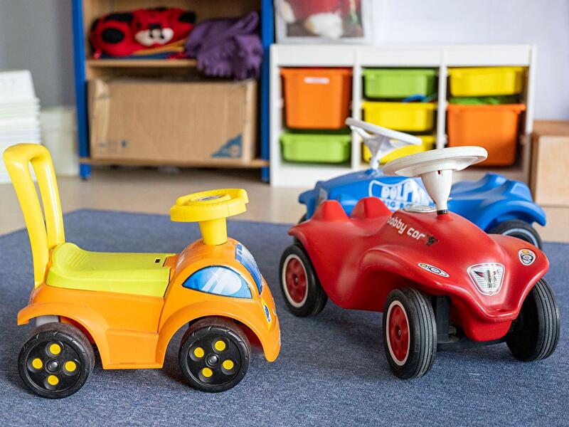 Bunte Bobby-Cars und Rutschautos stehen auf einem blauen Teppich in einem Kita-Gruppenraum, im Hintergrund ein Regal mit Aufbewahrungsboxen.