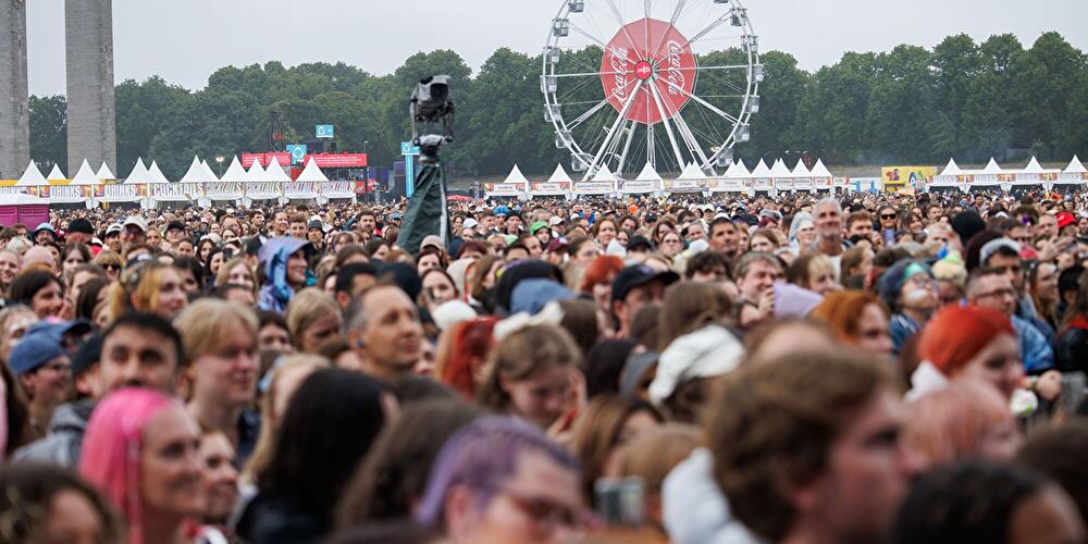 Dichte Menschenmenge auf dem Lollapalooza-Festivalgelände in Berlin, im Hintergrund ein Riesenrad, Verpflegungsstände und Bäume unter grauem Himmel.