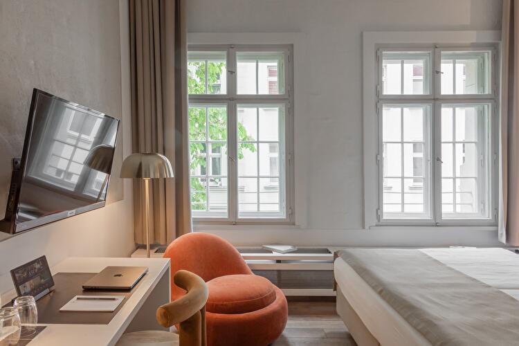 Hotelzimmer mit orangefarbenem Sessel, Schreibtisch mit Laptop und Wand-TV, im Hintergrund zwei große Sprossenfenster und ein Bett.