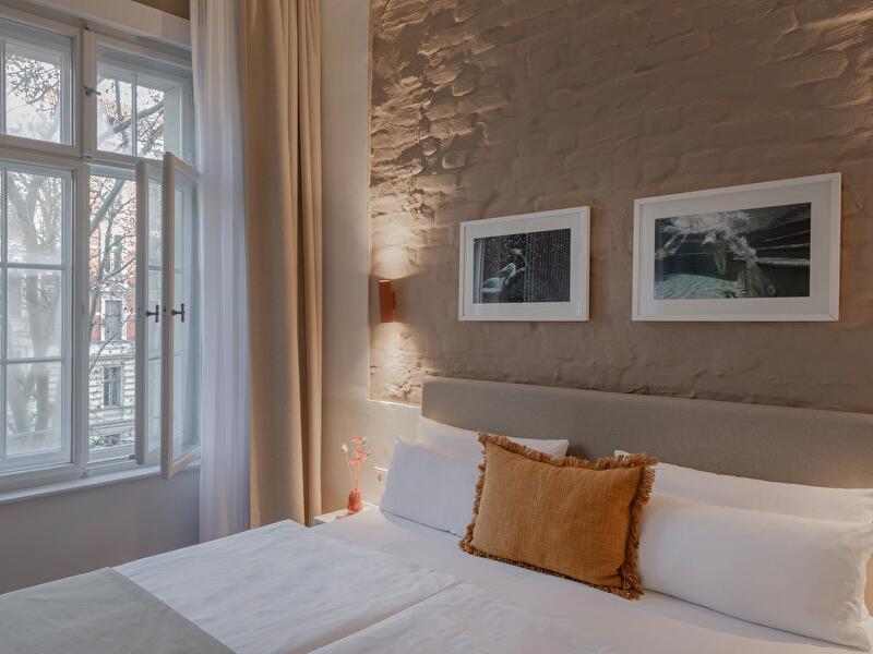 Hotelzimmer im Hotel Oderberger mit Doppelbett, orangefarbenem Zierkissen und zwei gerahmten Fotografien an einer rustikalen Wand, im Hintergrund ein offenes Fenster mit Blick auf Bäume.