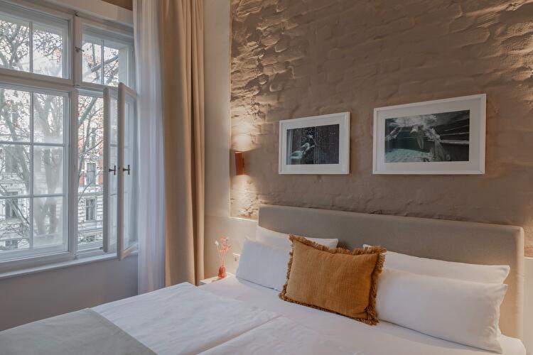 Hotelzimmer im Hotel Oderberger mit Doppelbett, orangefarbenem Zierkissen und zwei gerahmten Fotografien an einer rustikalen Wand, im Hintergrund ein offenes Fenster mit Blick auf Bäume.