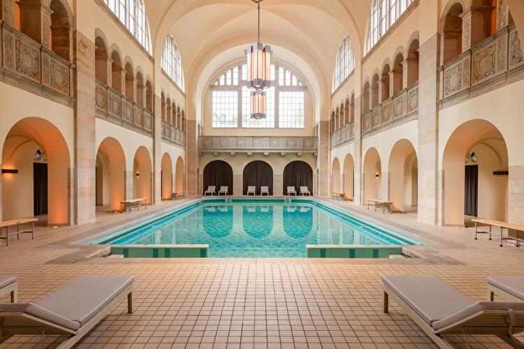Historisches Schwimmbad des Hotel Oderberger mit türkisfarbenem Becken unter einer Tonnengewölbedecke, umgeben von Arkadengängen und Liegestühlen.
