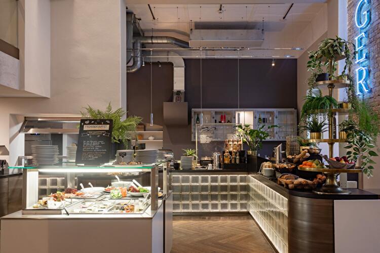 Frühstücksbuffet-Bereich mit Vitrine für kalte Speisen, Getränkestation und Obstschale, dekoriert mit Pflanzen und einer Neon-Schrift.