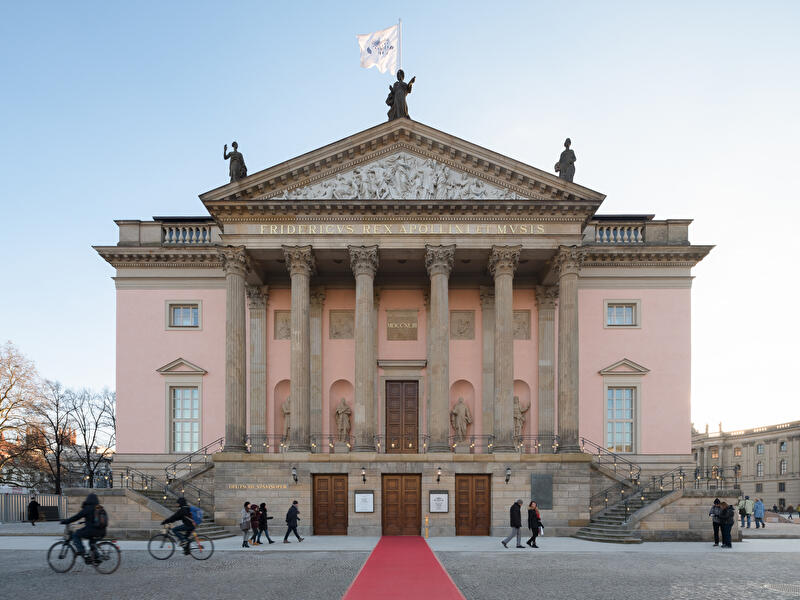Frontalansicht der Staatsoper Unter den Linden in Berlin mit Säulenfassade, Freitreppe und rotem Teppich. Vor dem Gebäude gehen Passanten und fahren Radfahrer.