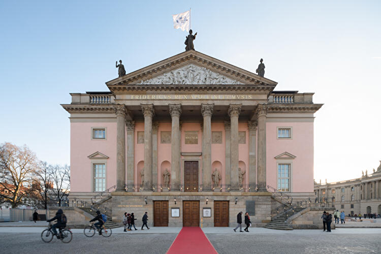 Frontalansicht der Staatsoper Unter den Linden in Berlin mit Säulenfassade, Freitreppe und rotem Teppich. Vor dem Gebäude gehen Passanten und fahren Radfahrer.