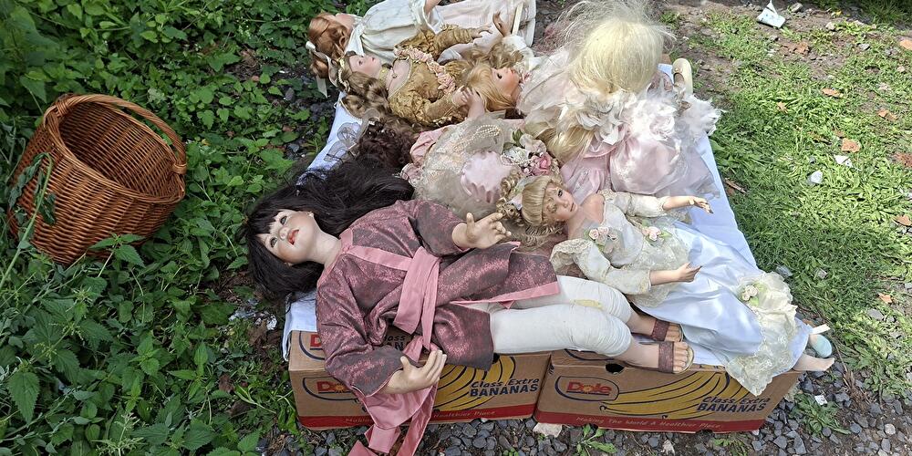 Auf dem Flohmarkt liegen mehrere Porzellanpuppen in aufwendigen Kleidern auf Kisten neben einem Korb im Gras.