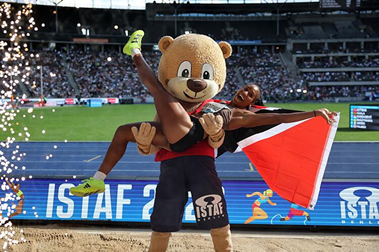 Athletin Tyra Gittens lässt sich lachend vom ISTAF-Bärenmaskottchen tragen, dabei hält sie eine Flagge von Trinidad und Tobago, umgeben von Funkenregen.