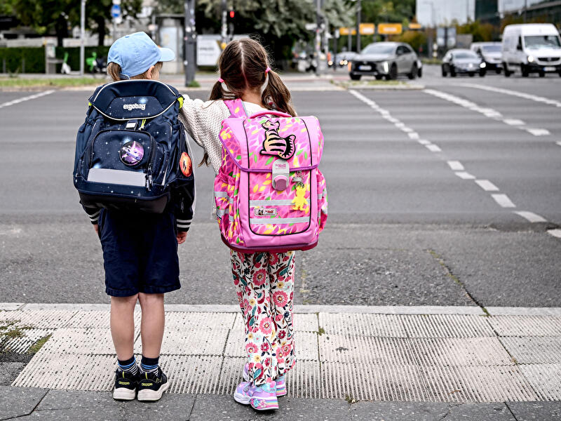 Zwei Kinder mit Schulranzen stehen von hinten an einer Straße und warten darauf, sie zu überqueren, im Hintergrund fließender Verkehr.
