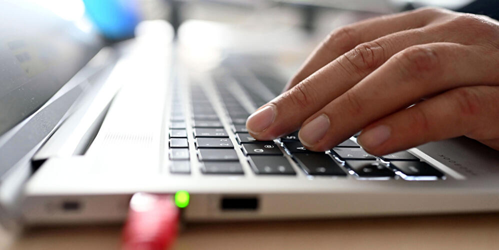 Hand tippt auf Laptop-Tastatur, Netzwerkanschluss mit Kabel im Vordergrund.