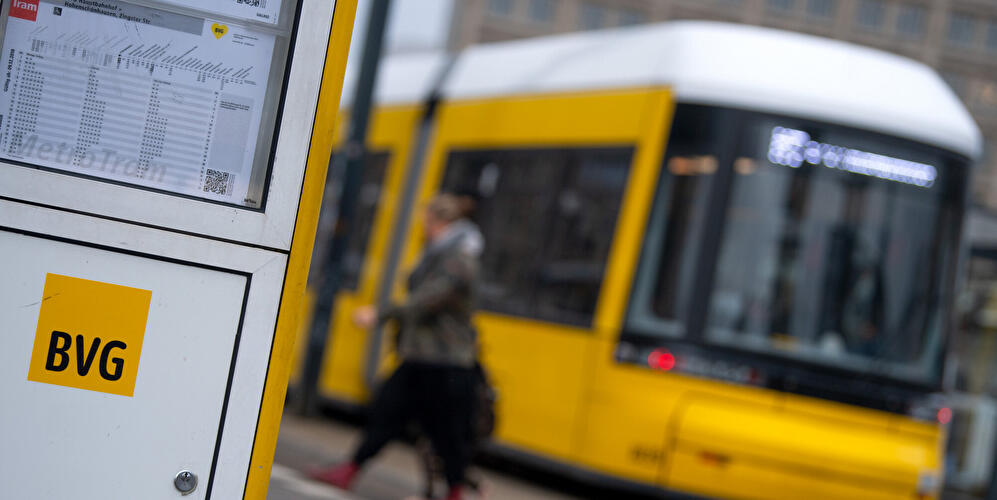 BVG-Fahrplan an Haltestelle, im Hintergrund eine gelbe Straßenbahn.