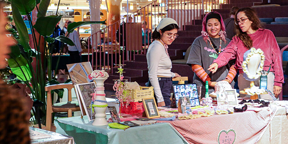 Besucher:innen betrachten handgefertigte Strick- und Kunstprodukte an einem Marktstand.
