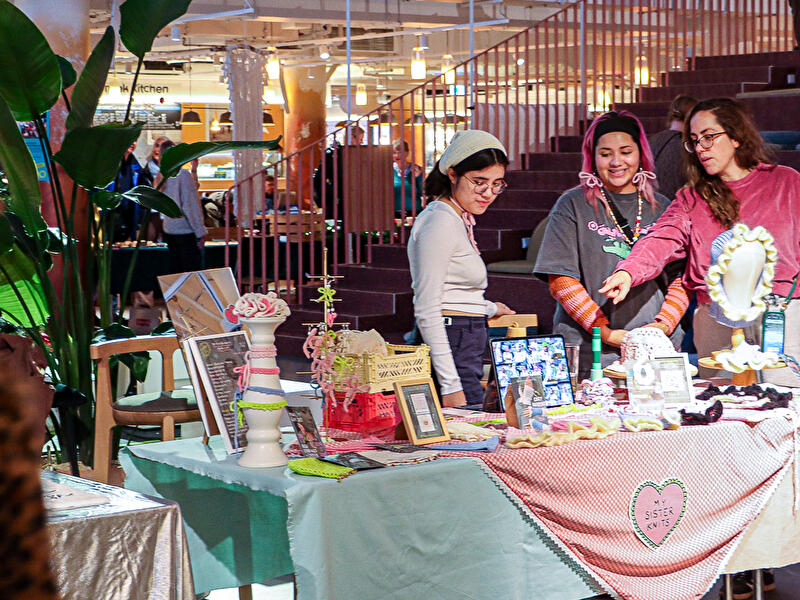 Besucher:innen betrachten handgefertigte Strick- und Kunstprodukte an einem Marktstand.