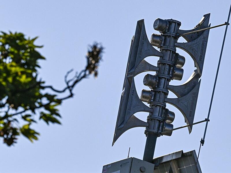 Sirenenmast mit mehreren Lautsprechern vor Himmel und Baumzweigen.