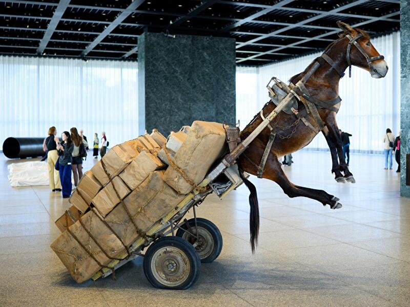 Lebensgroße Pferdeskulptur mit Warenkarren steigt auf den Hinterbeinen in der Halle der Neuen Nationalgalerie, umgeben von Besucher:innen.
