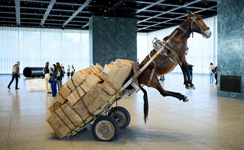 Lebensgroße Pferdeskulptur mit Warenkarren steigt auf den Hinterbeinen in der Halle der Neuen Nationalgalerie, umgeben von Besucher:innen.