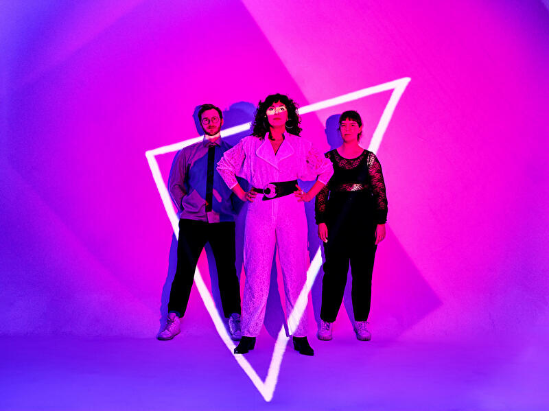 Drei Personen vor pink-lila Hintergrund, beleuchtet von einem weißen neonleuchtenden Dreieck.