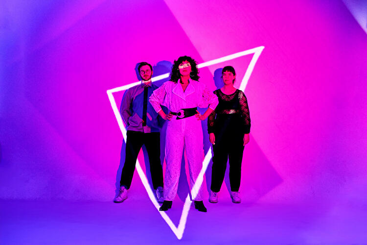 Drei Personen vor pink-lila Hintergrund, beleuchtet von einem weißen neonleuchtenden Dreieck.