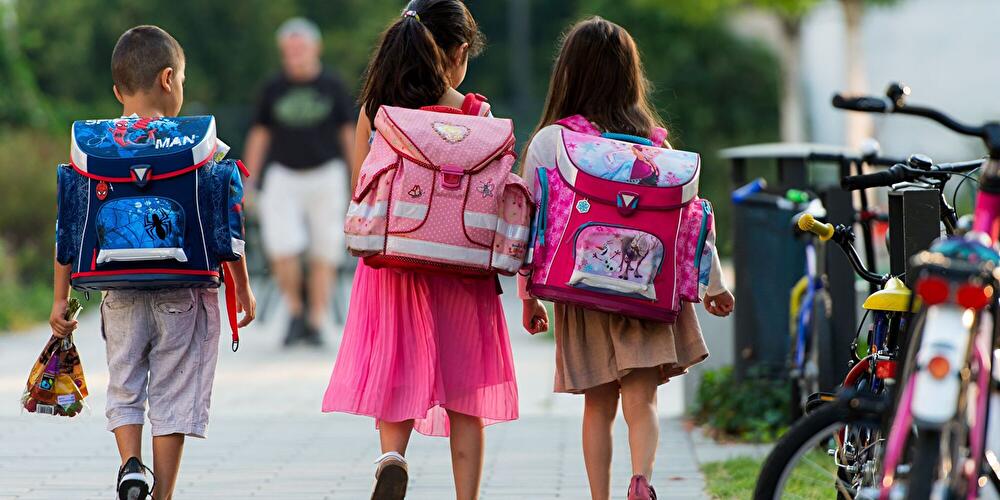 Drei Kinder mit Schulranzen laufen auf einem Gehweg zwischen Fahrrädern und Bäumen.