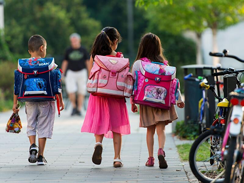 Drei Kinder mit Schulranzen laufen auf einem Gehweg zwischen Fahrrädern und Bäumen.