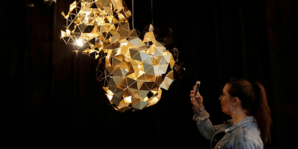 Person fotografiert eine leuchtende Lichtinstallation aus goldfarbenen, geometrischen Elementen in dunklem Raum.