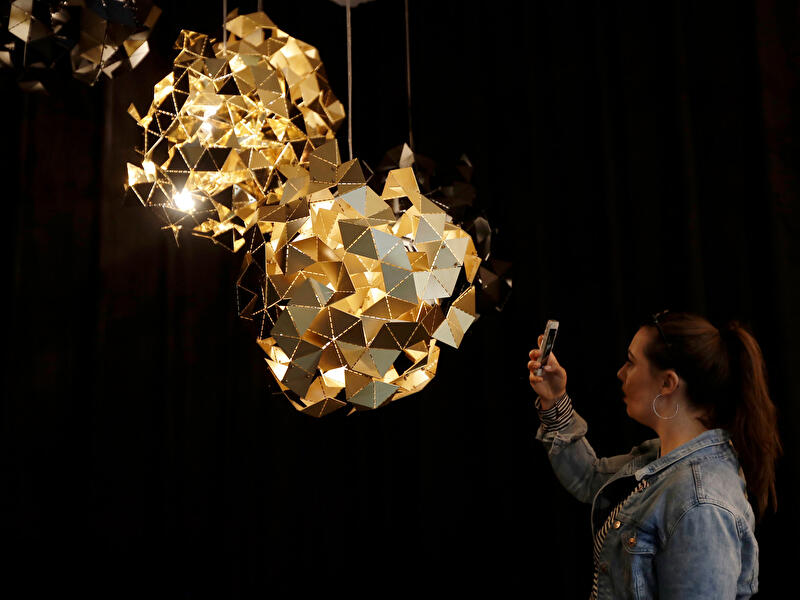 Person fotografiert eine leuchtende Lichtinstallation aus goldfarbenen, geometrischen Elementen in dunklem Raum.