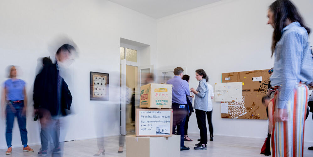 Besucher:innen bewegen sich durch einen hellen Galerieraum mit Kunstwerken und einer Installation aus Kartons.