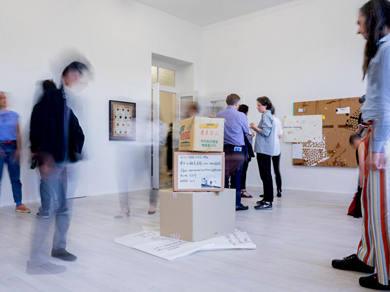 Besucher:innen bewegen sich durch einen hellen Galerieraum mit Kunstwerken und einer Installation aus Kartons.