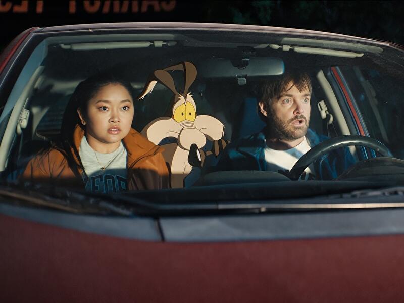 Zwei Personen sitzen mit der Comicfigur Wile E. Coyote in einem Auto und blicken aufmerksam durch die Frontscheibe.