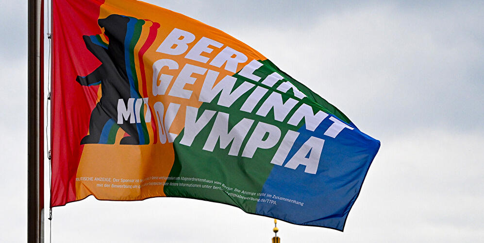 Regenbogenfarbene Fahne mit dem Schriftzug „Berlin gewinnt Olympia“ weht vor Berliner Kulisse.