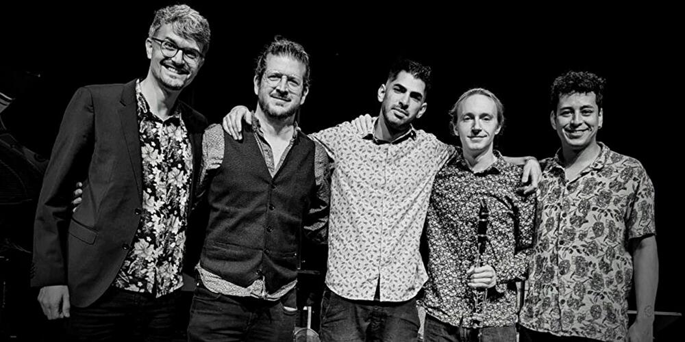 Fünf Musiker stehen eng beisammen auf einer Bühne und posieren nach einem Auftritt für ein Gruppenfoto.