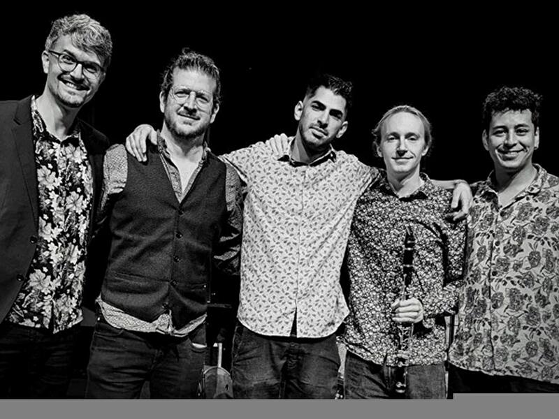 Fünf Musiker stehen eng beisammen auf einer Bühne und posieren nach einem Auftritt für ein Gruppenfoto.