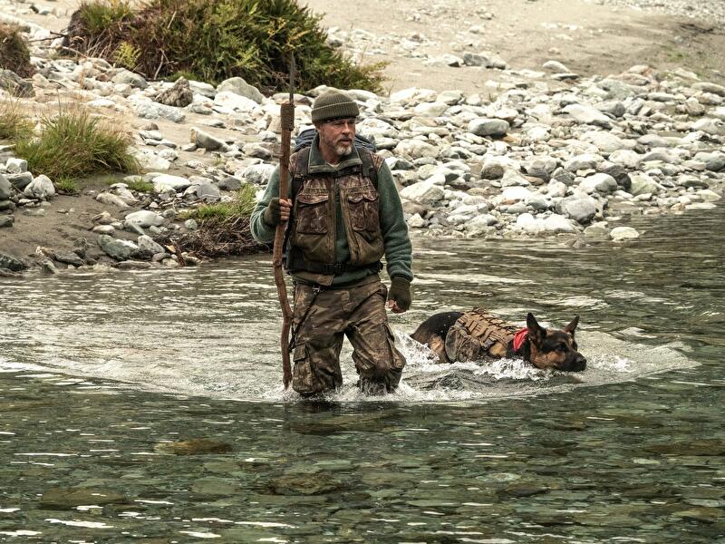 Person und ausgerüsteter Hund durchqueren einen flachen Fluss in einer steinigen Wildnis.
