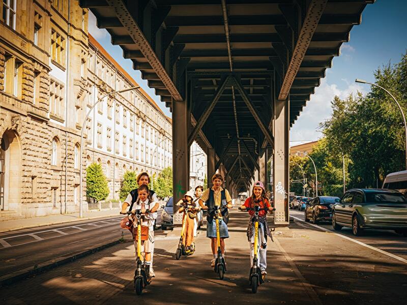Mehrere Kinder fahren mit E-Scootern unter einer Hochbahntrasse durch eine Berliner Straße mit parkenden Autos.